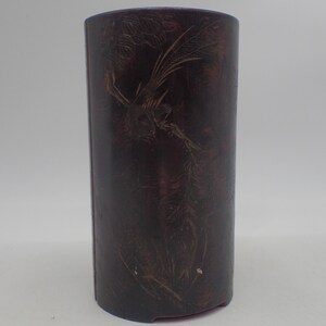 May include: A dark brown cylindrical vase with a carved design of a bird in flight. The vase has a smooth, polished finish.