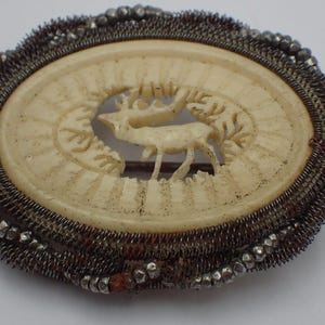 Può includere: Spilla ovale antica con un centro color crema raffigurante un cervo intagliato. La spilla è incorniciata da un bordo dettagliato in filo e perline nei toni dell'argento e del marrone, con chiusura a spillo.