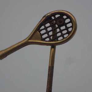 Peut inclure: Une épingle en métal de couleur or en forme de raquette de tennis. La raquette est détaillée avec un motif en maille et une petite balle au centre. L'épingle est fixée à une fine tige en métal de couleur or.