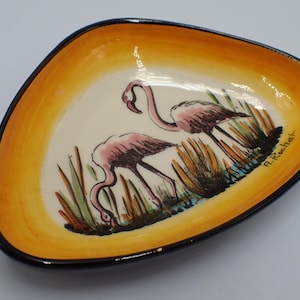 May include: Triangular ceramic dish with a hand-painted design of two pink flamingos in a marsh setting. The dish has a yellow and black border, and the artist's signature, "A. Rechat," is visible.