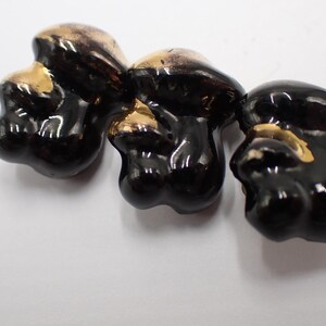 Può includere: Tre oggetti lucidi in ceramica nera a forma di fungo con accenti dorati. I funghi hanno una forma liscia e arrotondata e sono disposti su una superficie bianca. I riflessi dorati aggiungono un tocco di eleganza alle forme scure e organiche.
