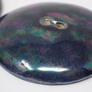 Pu&ograve; includere: Primo piano di un bottone in ceramica blu scuro iridescente con un foro al centro.
