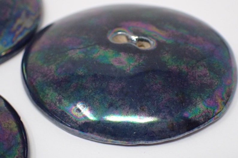 Pu&ograve; includere: Primo piano di un bottone in ceramica blu scuro iridescente con un foro al centro.