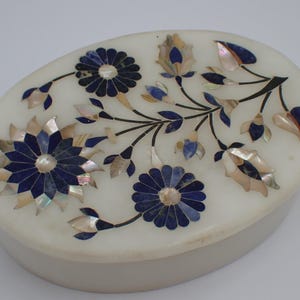 Op de afbeelding: Ovale witte marmeren doos met een bloemmotief. De bloemen zijn gemaakt van donkerblauwe en iriserende schelpen. De doos heeft een lichtgrijze basis en een wit deksel. Het bloemmotief bevat donkere stelen en bladeren.