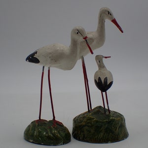 Peut inclure: Trois figurines de cigognes blanches avec des pattes rouges et des marques noires. Les cigognes sont debout sur des socles verts.