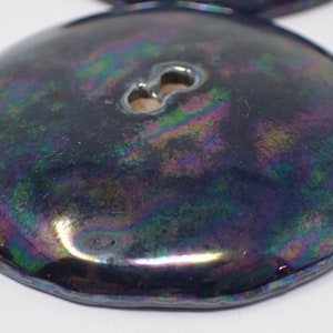 Pu&ograve; includere: Un bottone rotondo nero iridescente con un gambo a due fori. Il bottone ha una finitura liscia e lucida e un bordo leggermente rialzato.