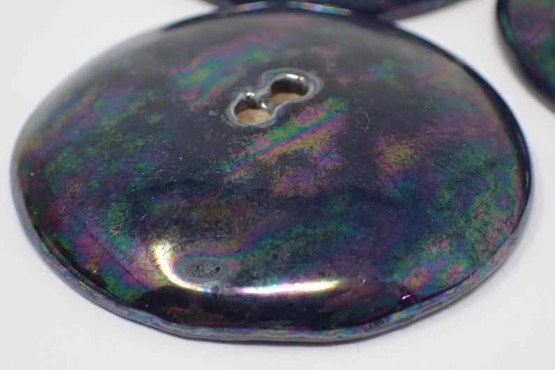 Pu&ograve; includere: Un bottone rotondo nero iridescente con un gambo a due fori. Il bottone ha una finitura liscia e lucida e un bordo leggermente rialzato.