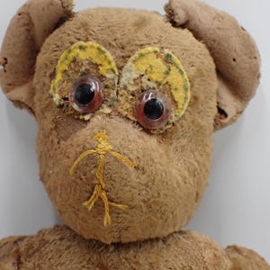 Op de afbeelding: Een vintage teddybeer met bruine vacht, grote plastic ogen en gele en groene accenten rond de ogen. De beer heeft een gestikte gele mond en zichtbare slijtage, waardoor hij een geliefde uitstraling heeft.