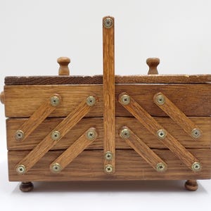 Peut inclure: Une boîte à couture en bois brun moyen dotée d'un design de rangement à plusieurs niveaux en accordéon. La boîte comprend une poignée verticale centrale, deux petits boutons en bois sur les couvercles et quatre petits pieds, fixés par des vis.