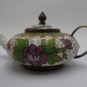 Può includere: Una piccola teiera bianca con un motivo floreale. La teiera è decorata con foglie verdi, fiori viola e dettagli dorati. La teiera ha un piccolo beccuccio e un manico.