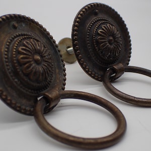 Op de afbeelding: Twee antieke messing deurknopjes met sierlijke bloemmotieven en ringgrepen.