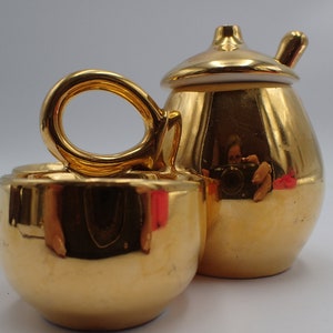 以下が含まれることがあります： 金メッキのセラミック製のティーセットで、ティーポットとカップがセットになっています。ティーポットは、小さな取っ手と注ぎ口が付いた蓋が付いています。カップは取っ手があり、丸みを帯びた形をしています。
