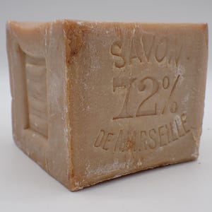 Op de afbeelding: Een rechthoekig blok beige zeep met de tekst "SAVON 72% DE MARSEILLE" in het oppervlak gegraveerd. De zeep heeft een rustieke uitstraling en een uitgesneden gedeelte aan één kant.