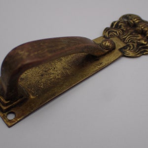 Op de afbeelding: Een messing deurgreep met een decoratief, sierlijk ontwerp. De greep heeft een gebogen, lusvormige vorm en een rechthoekige basis.