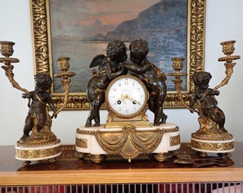 May include: An antique clock with two cherub figures and two candelabras. The clock has a white face with black Roman numerals and gold accents. The cherubs are bronze-colored, and the candelabras are gold-toned.