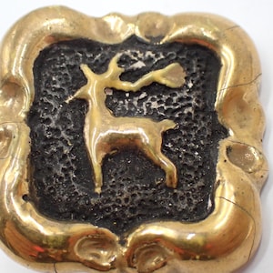 May include: A square brass button featuring a raised, polished gold-tone deer silhouette centered against a dark, textured, recessed background. The button has a smooth, rounded, and slightly irregular gold-tone border with visible surface cracks.