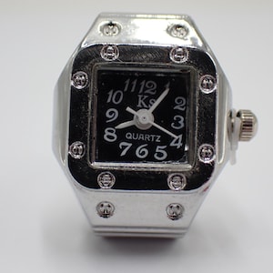May include: Silver ring watch with a black square face. The watch face has white numerals and hands, and the word "QUARTZ" is at the bottom. The ring is rectangular with decorative rivets around the face.