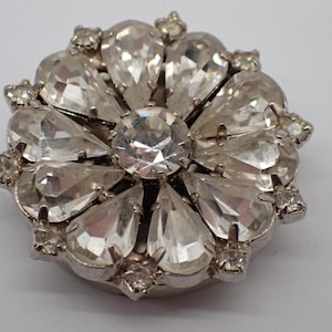 Op de afbeelding: Zilverkleurige broche met een bloemmotief. De broche heeft een grote, heldere, ronde middensteen, omringd door druppelvormige heldere stenen. Kleinere heldere stenen accentueren de buitenste randen.