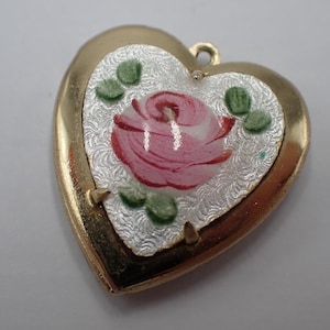 May include: A heart-shaped gold-tone pendant featuring a central white textured inlay with a painted pink rose and small green leaves. The pendant has a small metal loop at the top for attachment and a smooth, polished gold-colored metal border.