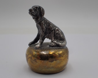 Pode incluir: Uma figura de cão prateada sentada em uma base dourada. O cão está olhando para a esquerda e tem uma coleira ao redor do pescoço.