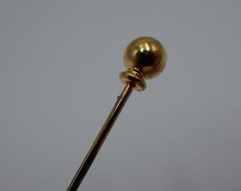 Op de afbeelding: Een goudkleurige hoedenspeld met een bolvormige top. De speld heeft een lange, slanke schacht en een decoratieve, afgeronde basis onder de bol. De speld staat tegen een effen, lichtgrijze achtergrond.