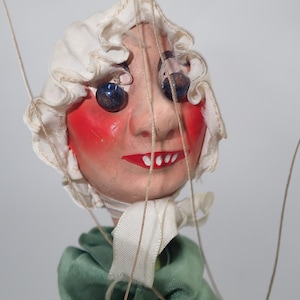 Pode incluir: Uma cabeça de marioneta vintage com um rosto pintado, bochechas rosadas e uma boca vermelha. A marioneta usa um gorro branco com acabamento em babados e uma roupa verde. Os olhos são grandes e azul escuro. Fios são anexados para manipulação.
