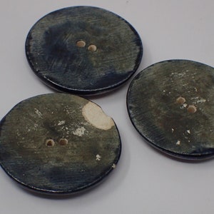 Pu&ograve; includere: Tre bottoni rotondi in ceramica blu con due fori ciascuno. I bottoni hanno un aspetto screziato e invecchiato.