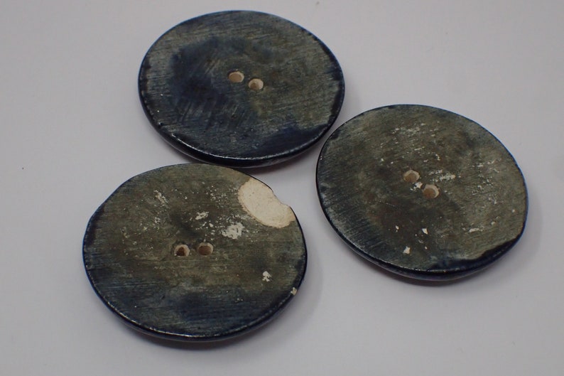 Pu&ograve; includere: Tre bottoni rotondi in ceramica blu con due fori ciascuno. I bottoni hanno un aspetto screziato e invecchiato.