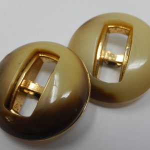 Puede incluir: Dos botones redondos de plástico de color marrón y crema con un bucle de metal dorado. Los botones tienen un acabado liso y brillante.
