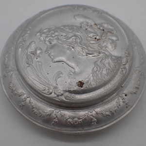 Peut inclure: Un couvercle rond en métal argenté avec un motif en relief. L'image centrale est un profil de femme aux cheveux flottants, entourée d'éléments floraux et décoratifs. Le couvercle a un bord surélevé avec des détails en relief supplémentaires.