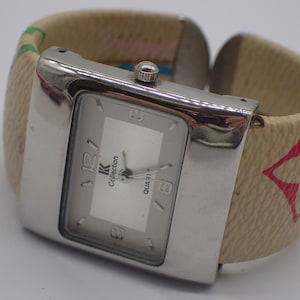 Può includere: Un orologio argentato con quadrante bianco e lancette nere. L'orologio ha un cinturino in pelle marrone con un motivo rosso e bianco. Il quadrante dell'orologio dice "K Collection Quartz".