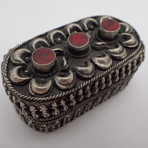Può includere: Una piccola scatola ovale in argento con intricati disegni floreali e due accenti di pietre preziose rosse.
