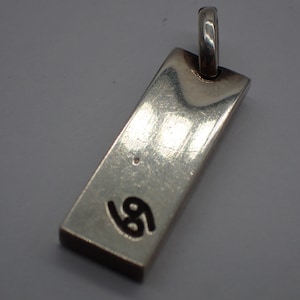 Puede incluir: Un colgante de plata con forma rectangular y el número "69" grabado en negro en la parte delantera.