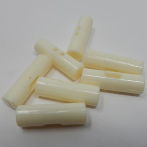 Puede incluir: Seis botones de plástico de color blanco roto con dos agujeros cada uno. Los botones están dispuestos en una pila.