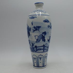 Peut inclure: Un vase en céramique blanc et haut, orné d'illustrations bleues peintes à la main. Le vase présente un col étroit et un corps plus large, avec une scène détaillée de personnages et une clôture. La base a un motif décoratif.