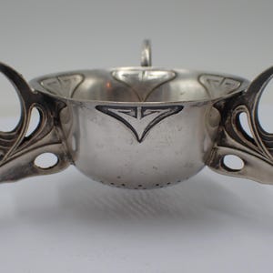 Op de afbeelding: Een zilverkleurige Art Nouveau theezeef met een klein handvat en twee grote handvatten. De zeef heeft drie poten en decoratieve bloemmotieven. De kom heeft een klein handvat en decoratieve elementen.