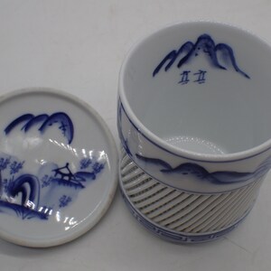 以下が含まれることがあります： 青と白の山の風景のデザインが施された、白磁の茶碗と蓋。茶碗の底には格子模様があり、内側には「五五」の文字が書かれている。