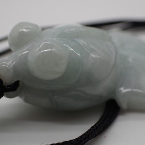 Op de afbeelding: Lichtgroene jade hanger in de vorm van een vis. De jade heeft een glad, gepolijst oppervlak en een doorschijnend uiterlijk. Het visontwerp is gedetailleerd en elegant. De hanger is bevestigd aan een zwart koord.