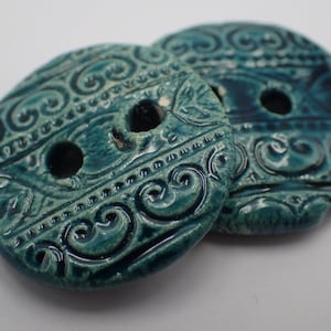 Puede incluir: Dos botones de cerámica azul verdoso con intrincados diseños de espirales y flores. Los botones tienen dos agujeros cada uno.
