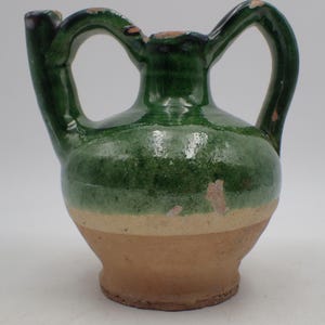 Può includere: Una piccola brocca in ceramica fatta a mano con una combinazione di colori verde e beige. La brocca presenta un corpo arrotondato, un beccuccio e due manici ad arco. La base è beige chiaro, con una fascia verde al centro e una parte superiore verde.