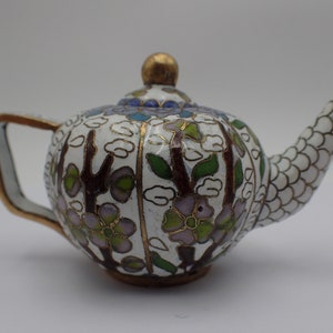 Può includere: Una piccola teiera bianca con un motivo floreale in tonalità rosa, verde e marrone. La teiera ha un coperchio dorato e un beccuccio con un motivo a scaglie bianco e dorato.