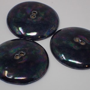 Op de afbeelding: Drie iriserende zwarte knopen met elk één gat. De knopen hebben een glad, afgerond oppervlak en een licht verhoogde rand.