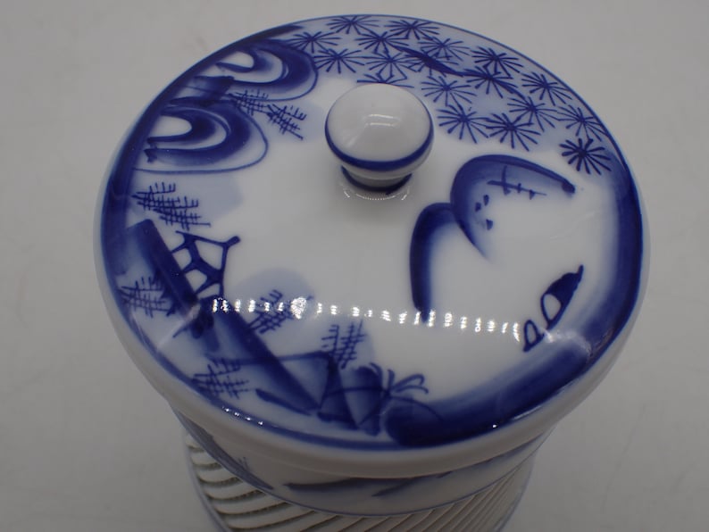 以下が含まれることがあります： 青と白のフラワーデザインが施された白い陶器の壺。蓋の上にはノブがあり、青と白のフラワーデザインが施されています。壺の底にはリブ模様があります。