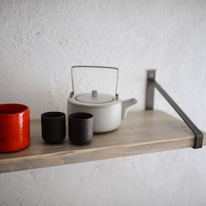 May include: A red ceramic mug, two small dark brown cups, and a white teapot with a metal handle sit on a wooden shelf. The shelf is supported by a black metal bracket against a textured white wall.