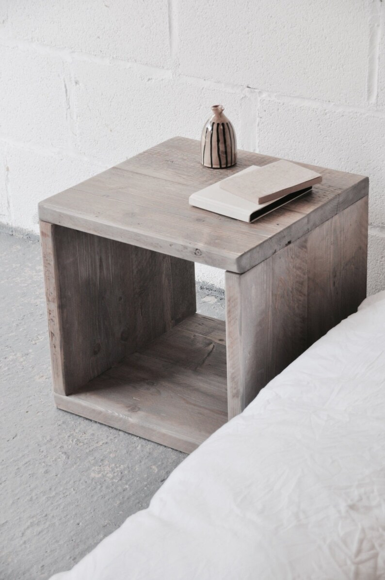 May include: A rustic wooden end table with a single shelf. The table is made of reclaimed wood and has a natural gray finish. A small ceramic vase with black stripes and a book are on top of the table.