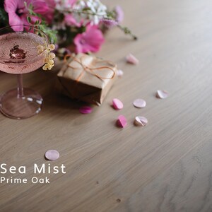 Puede incluir: Naturaleza muerta rom&aacute;ntica con un c&oacute;ctel rosa en una copa, un regalo envuelto y p&eacute;talos esparcidos. Flores rosas y el texto "Sea Mist Prime Oak" en el fondo. La imagen tiene una paleta de colores suaves.
