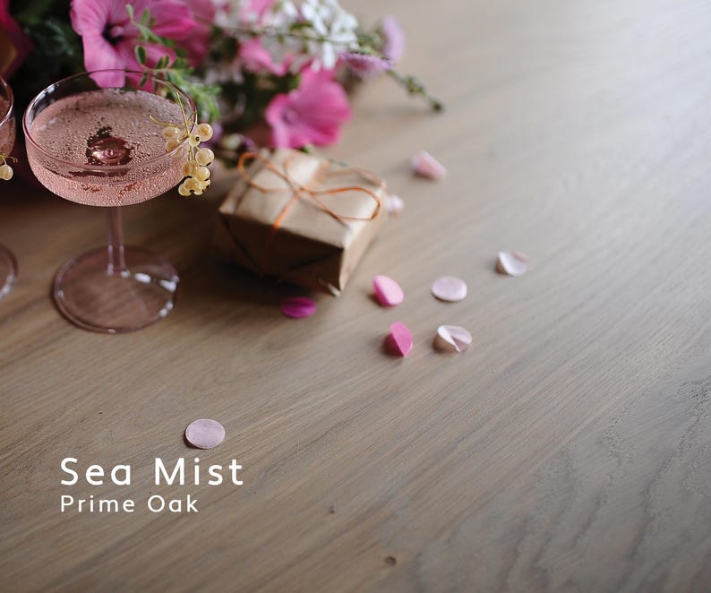 Puede incluir: Naturaleza muerta rom&aacute;ntica con un c&oacute;ctel rosa en una copa, un regalo envuelto y p&eacute;talos esparcidos. Flores rosas y el texto "Sea Mist Prime Oak" en el fondo. La imagen tiene una paleta de colores suaves.