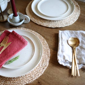 May include: A table setting with white speckled plates, gold cutlery, and a red napkin. The plates rest on woven placemats. A white candle holder with a burgundy candle and gold spoons are also visible. The setting is on a wooden table.