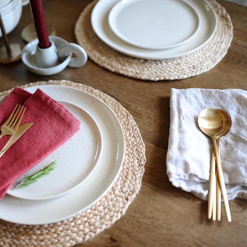 May include: A table setting with white speckled plates, gold cutlery, and a red napkin. The plates rest on woven placemats. A white candle holder with a burgundy candle and gold spoons are also visible. The setting is on a wooden table.
