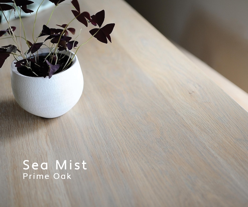 May include: A light-toned oak wood surface with a visible grain pattern, labeled Sea Mist Prime Oak. A small, round, white textured ceramic pot contains a plant with dark purple, triangular-shaped leaves and thin, light green stems.